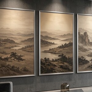 May include: A triptych of three framed landscape paintings featuring a sepia-toned mountain range, rolling hills, and a winding river. The artwork displays a misty, atmospheric valley with scattered trees and rocky terrain, all enclosed in thin silver frames.