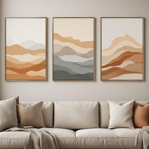 Peut inclure: Un ensemble de trois impressions d'art mural rectangulaires verticales représentant des paysages de montagnes abstraits en couches dans des tons beige, ocre, terre cuite et gris, encadrées par un fin cadre doré et alignées horizontalement.