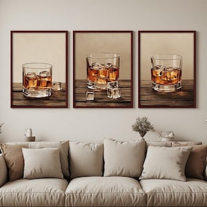 May include: A set of three framed wall art prints, each featuring a clear glass tumbler filled with amber-colored liquid and ice cubes resting on a dark wooden surface. The frames are a deep brown color, and the prints have a neutral background.