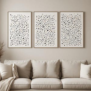 Può includere: Un set di tre stampe artistiche rettangolari da parete con un motivo di punti neri e oro metallizzato sparsi su sfondo bianco. Ogni stampa è racchiusa in una sottile cornice di legno chiaro, disposte orizzontalmente in fila.
