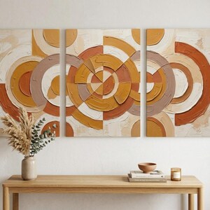 Peut inclure: Un ensemble d'art mural en trois panneaux présentant un motif géométrique texturé de cercles et d'arcs superposés dans des tons orange brûlé, jaune moutarde, crème et brun sourd. L'application épaisse de peinture crée une surface tactile.