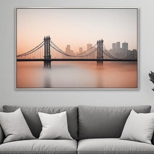 Peut inclure: Une impression murale rectangulaire représentant un pont suspendu au-dessus d'un plan d'eau avec une ligne d'horizon urbaine en arrière-plan. La scène est capturée dans des tons doux de pêche et d'orange avec un effet de brouillard atmosphérique.