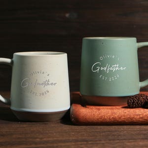 May include: Two ceramic mugs with a speckled finish. The left mug is off-white and features the text "Olivia's Godmother EST. 2026". The right mug is sage green and features the text "Olivia's Godfather EST. 2026". Both mugs have a smooth handle.