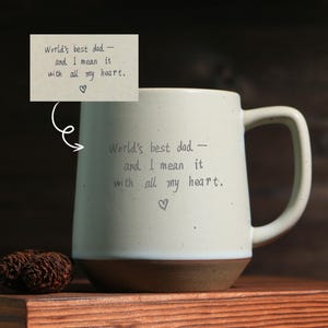 May include: Cream-colored ceramic mug with a brown base and a large handle. The mug is inscribed with the words "World's best dad - and I mean it with all my heart." A small heart symbol is below the text.