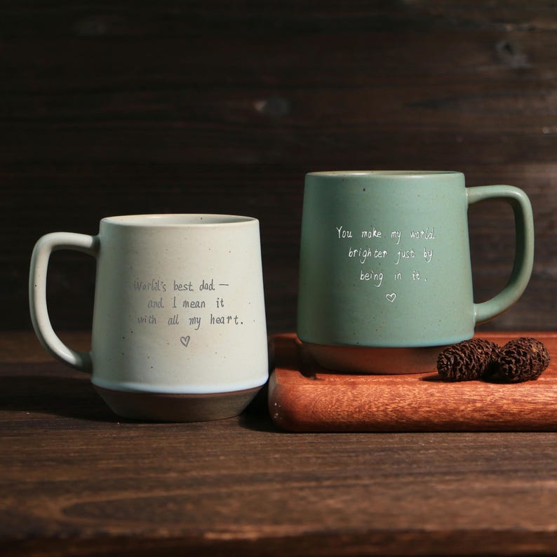 May include: Two ceramic mugs with messages. One is off-white with the text "World's best dad - and I mean it with all my heart." The other is green with the text "You make my world brighter just by being in it."