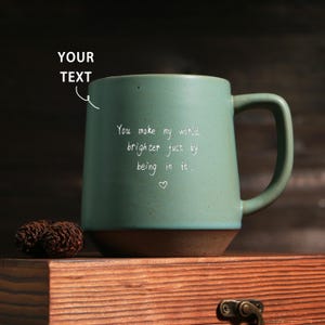 May include: A teal ceramic mug with a brown base and handle. The mug has the text "You make my world brighter just by being in it." written in white script. A small heart is below the text. The mug is on a wooden surface.