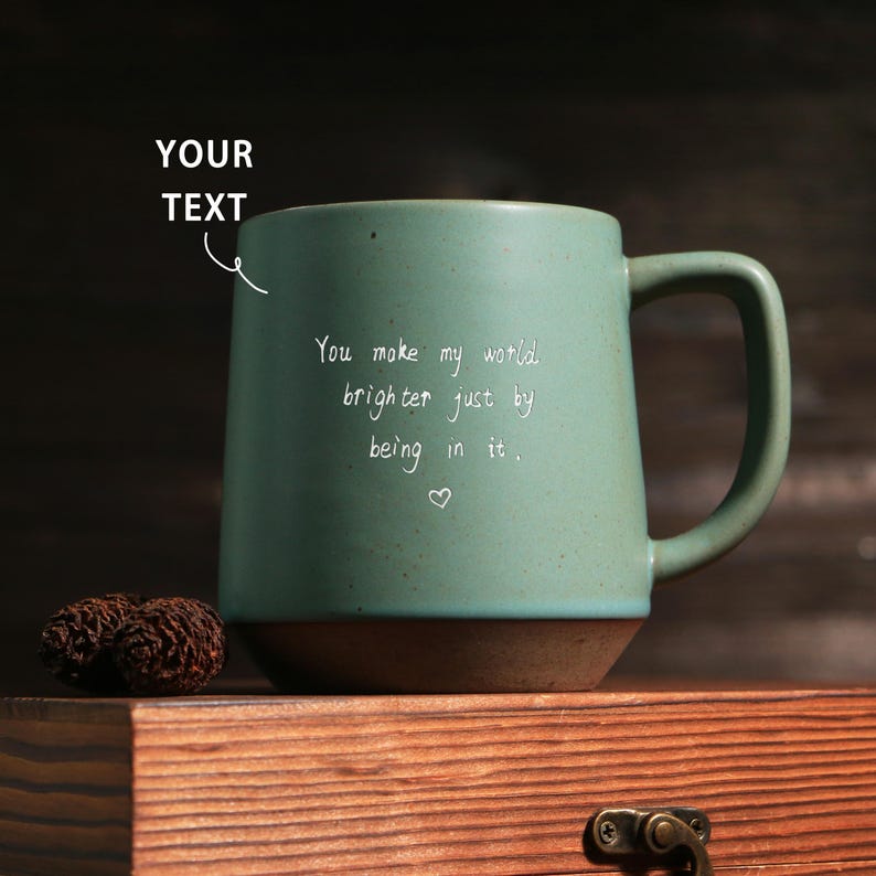 May include: A teal ceramic mug with a brown base and handle. The mug has the text "You make my world brighter just by being in it." written in white script. A small heart is below the text. The mug is on a wooden surface.