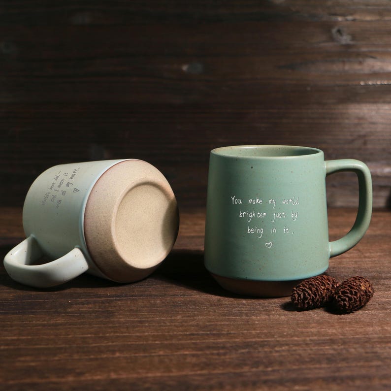 May include: Two ceramic mugs with a green exterior and brown base sit on a wooden surface. One mug is upright, displaying the text "You make my world brighter just by being in it." The other mug is on its side.