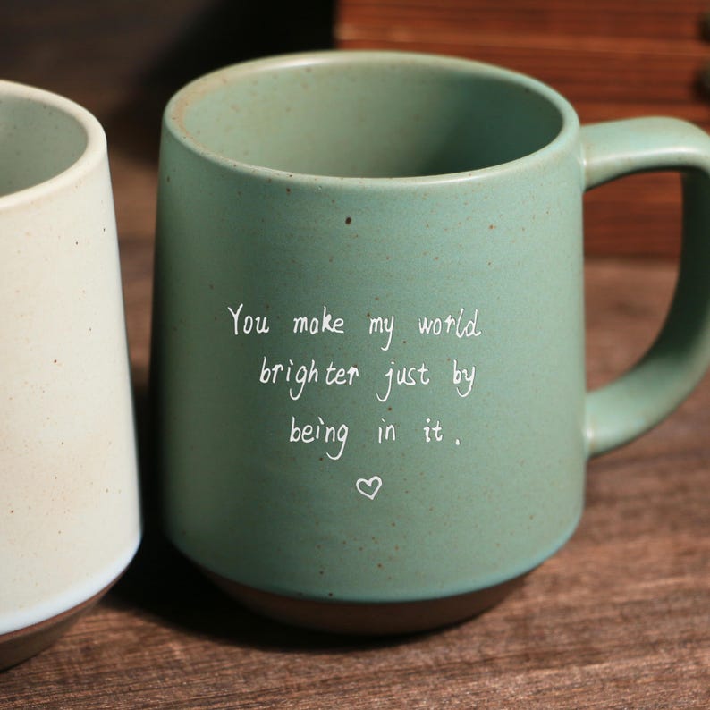 May include: A green ceramic mug with a brown base and a handle. The mug has the text "You make my world brighter just by being in it" in white script. A heart symbol is below the text.