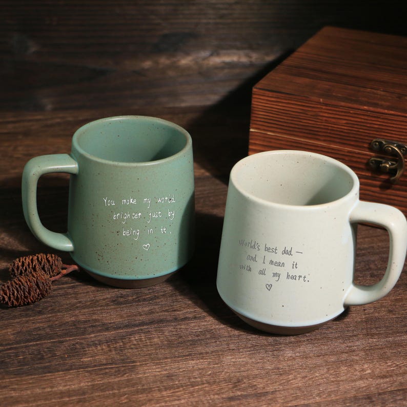 May include: Two ceramic mugs with handles. One is teal with the text "You make my world brighter just by being in it." The other is off-white with "World's best dad - and I mean it with all my heart."