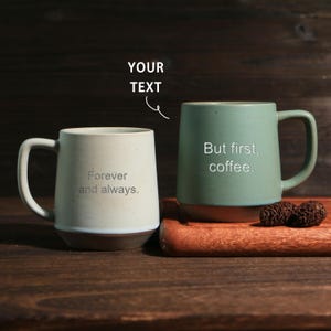 May include: Two ceramic coffee mugs. One is off-white with the text "Forever and always." The other is green with the text "But first, coffee." Both mugs have a handle and sit on a wooden surface.