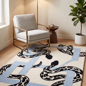 May include: A rectangular area rug featuring a cream-coloured background with a repeating pattern of black and white spotted snakes intertwined with a light blue geometric border. The design covers the entire surface of the floor covering.
