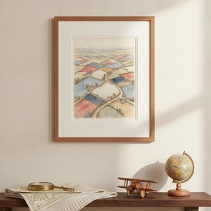 May include: A framed watercolor painting depicting an aerial view of a patchwork landscape with winding roads, fields in shades of blue, red, and tan, and small clusters of trees, enclosed in a light wood frame with a white mat board border.