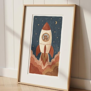 May include: Framed art print showing a rocket ship taking off into a starry night sky. The rocket is white and red, with a bear in the window. The artwork features a crescent moon and clouds in shades of brown and orange.