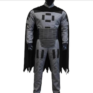 May include: A gray long-sleeved jumpsuit featuring a black high collar, a black cape with a jagged hem, and a wide black waist belt. The garment is decorated with black rectangular patches on the chest and thighs, and includes black wrist cuffs.