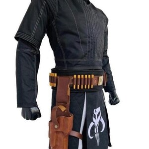 May include: A black long-sleeved tunic with white stitching, a high collar, and matching black pants. A brown leather belt features gold-toned ammunition loops and a holster. A white Mythosaur skull emblem is printed on the black fabric panel.