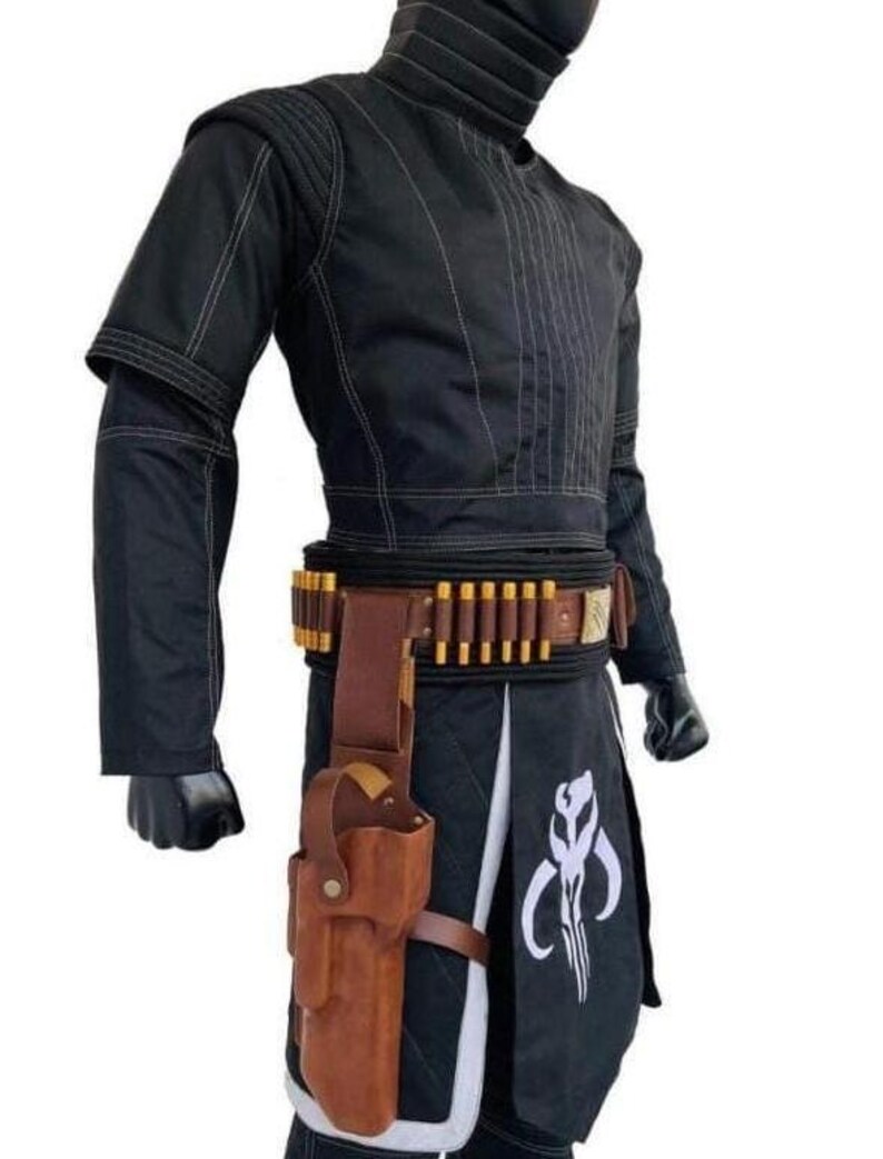 May include: A black long-sleeved tunic with white stitching, a high collar, and matching black pants. A brown leather belt features gold-toned ammunition loops and a holster. A white Mythosaur skull emblem is printed on the black fabric panel.