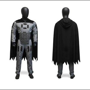 May include: A costume set consisting of a gray long-sleeved top and matching pants featuring black rectangular accents on the chest, shoulders, and thighs. A black hooded cape with a jagged hemline is worn over the outfit, which is displayed on a mannequin.