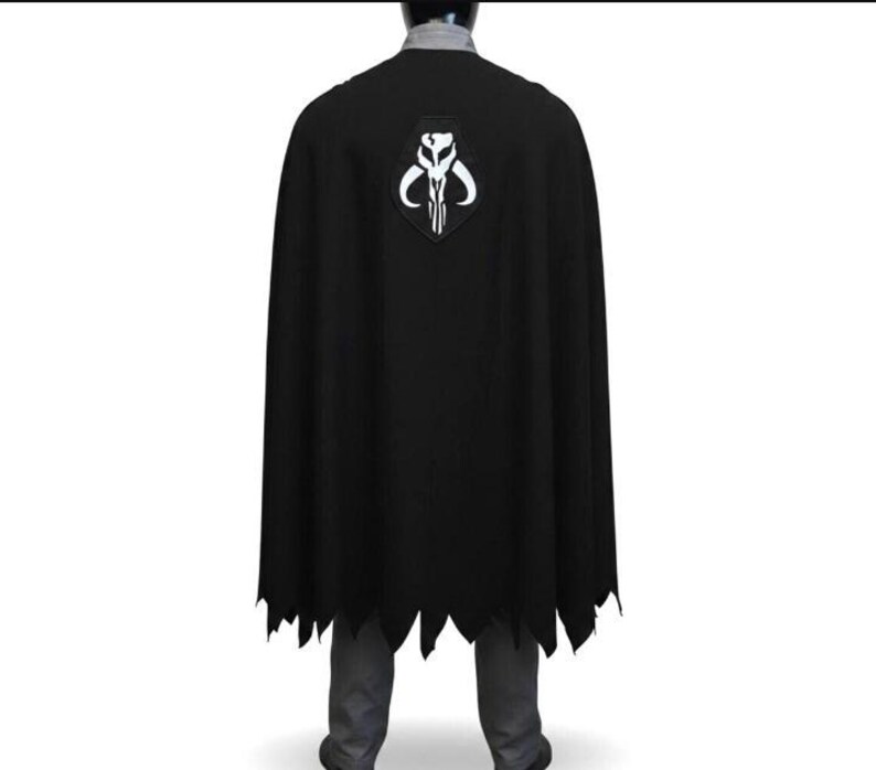 May include: A black cape featuring a white Mythosaur skull emblem centered on the upper back. The cape has a jagged, uneven hemline at the bottom and is worn over a grey collared shirt and grey trousers, covering the back of the torso.