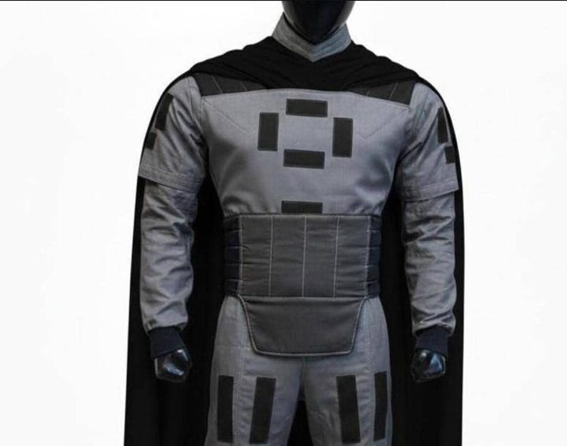 May include: A gray long-sleeved tunic and pants set featuring black rectangular patches on the chest, shoulders, and legs. The outfit includes a wide, horizontally stitched black waist belt and a long black cape attached at the shoulders.