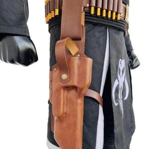 May include: A brown leather holster attached to a belt featuring a row of gold-colored cylindrical cartridges. The holster is worn over a black quilted garment with a white trim and a white graphic symbol printed on the front panel of the fabric.