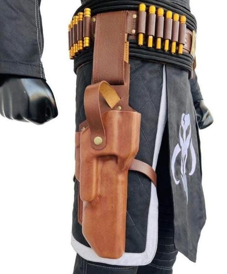 May include: A brown leather holster attached to a belt featuring a row of gold-colored cylindrical cartridges. The holster is worn over a black quilted garment with a white trim and a white graphic symbol printed on the front panel of the fabric.