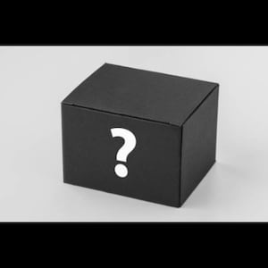 May include: A black, square box with a white question mark on the front. The box is sitting on a light gray surface. The box appears to be a gift box or a mystery item.