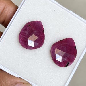 Puede incluir: Dos rubíes sueltos en forma de pera con un tono rojo magenta intenso y una superficie facetada. Cada piedra preciosa presenta una sección superior plana y pulida con un corte geométrico de múltiples facetas, sobre una bandeja blanca.