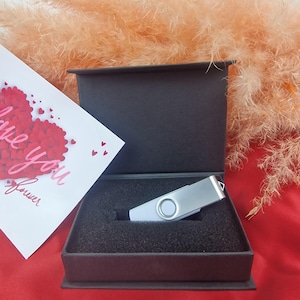 May include: A silver USB flash drive presented in a black gift box with a foam insert. A greeting card with a heart design and the words "love you forever" is visible. The background features dried ornamental grass.