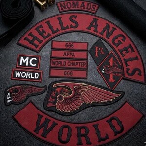May include: A collection of embroidered patches featuring the text HELLS ANGELS, NOMADS, MC, WORLD, AFFA, 666, and WORLD CHAPTER. The set includes a large winged skull emblem, a diamond-shaped 1% patch, and various rectangular and curved red and black labels.