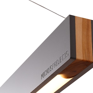 May include: A modern, rectangular pendant light fixture with a gray metal frame and a wood accent. The light fixture has a built-in LED light strip and the text "MICKUS PROJECTS" is engraved on the metal frame.