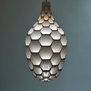 May include: A white pendant light with a geometric honeycomb design. The light is turned on and illuminates the room.
