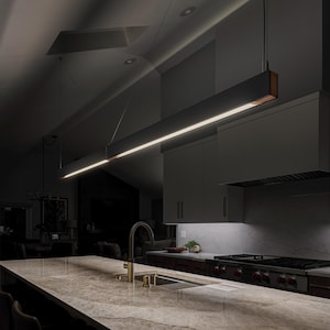 May include: A modern kitchen with a long, black, rectangular light fixture hanging over a kitchen island with a granite countertop. The light fixture has a copper trim and emits a warm white light.