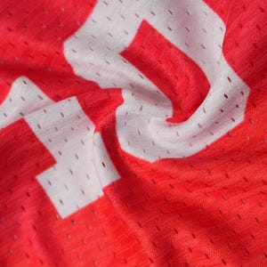 K&ouml;nnte beinhalten: Ein leuchtend rotes Sporttrikot aus atmungsaktivem Netzstoff mit kleinen, gleichm&auml;&szlig;ig verteilten Bel&uuml;ftungsl&ouml;chern. Das Kleidungsst&uuml;ck ist mit gro&szlig;en, kr&auml;ftigen wei&szlig;en Blockbuchstaben bedruckt, die direkt auf der strukturierten roten Oberfl&auml;che liegen.