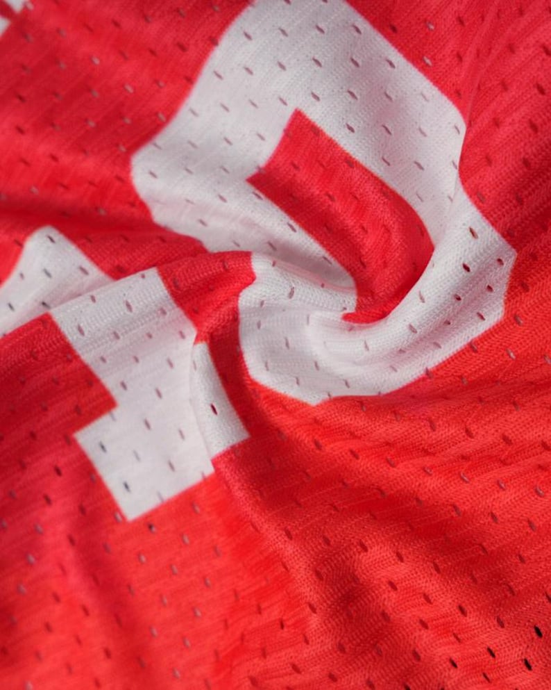 K&ouml;nnte beinhalten: Ein leuchtend rotes Sporttrikot aus atmungsaktivem Netzstoff mit kleinen, gleichm&auml;&szlig;ig verteilten Bel&uuml;ftungsl&ouml;chern. Das Kleidungsst&uuml;ck ist mit gro&szlig;en, kr&auml;ftigen wei&szlig;en Blockbuchstaben bedruckt, die direkt auf der strukturierten roten Oberfl&auml;che liegen.