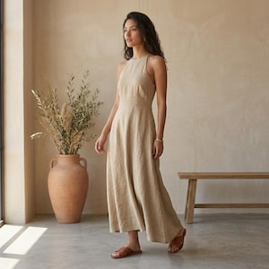 May include: A floor-length, sleeveless beige dress with a high neckline and a fitted bodice that flows into a relaxed, A-line skirt. The garment is paired with simple brown leather sandals, creating a cohesive and neutral-toned ensemble.
