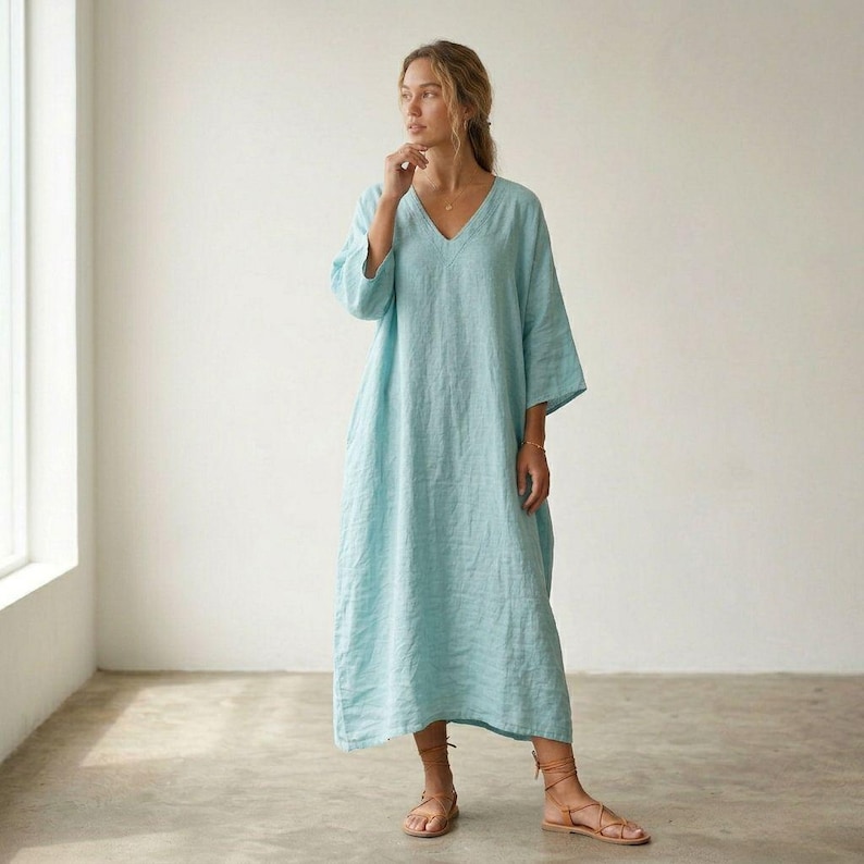 May include: A light blue, ankle-length dress with a V-neckline and three-quarter length sleeves. The garment features a loose, flowing silhouette and is crafted from a textured fabric. The dress is paired with brown strappy sandals.