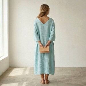 May include: A light blue midi-length dress with three-quarter length sleeves and a V-neckline on the back. The dress features a loose, flowing silhouette and is paired with brown strappy sandals and a small tan leather clutch held in the back.