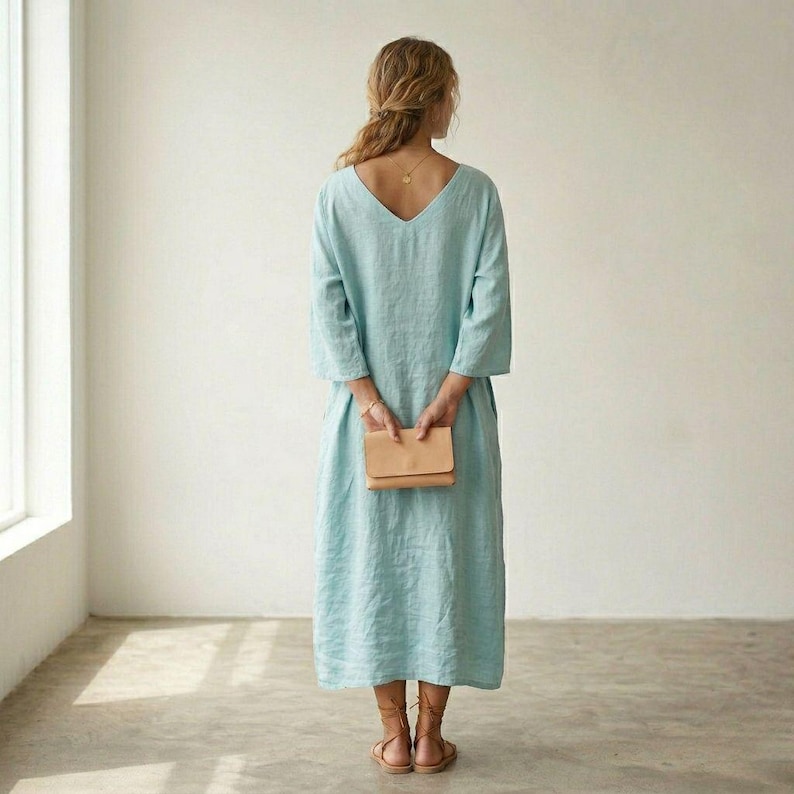 May include: A light blue midi-length dress with three-quarter length sleeves and a V-neckline on the back. The dress features a loose, flowing silhouette and is paired with brown strappy sandals and a small tan leather clutch held in the back.