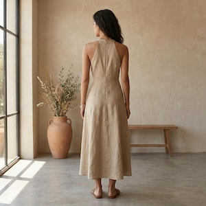 May include: A sleeveless midi dress in a solid beige color, featuring a high halter neckline, a fitted bodice, and a flared skirt that falls below the knees. The dress has a visible vertical seam running down the center back and a concealed zipper.