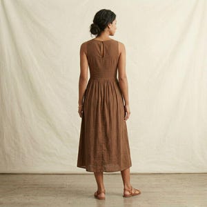 K&ouml;nnte beinhalten: Ein &auml;rmelloses, wadenlanges Kleid in einfarbigem Braun. Das Kleid hat ein eng anliegendes Oberteil mit Rundhalsausschnitt und einem Knopfverschluss am oberen R&uuml;cken, das in einen gerafften, weiten Rock &uuml;bergeht, der unter die Knie reicht.