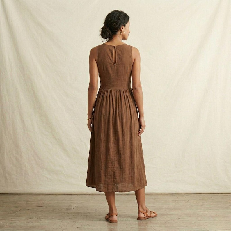 K&ouml;nnte beinhalten: Ein &auml;rmelloses, wadenlanges Kleid in einfarbigem Braun. Das Kleid hat ein eng anliegendes Oberteil mit Rundhalsausschnitt und einem Knopfverschluss am oberen R&uuml;cken, das in einen gerafften, weiten Rock &uuml;bergeht, der unter die Knie reicht.