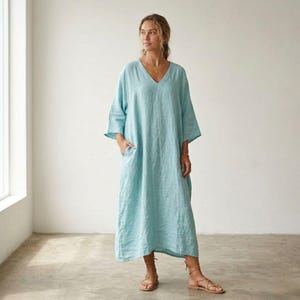 May include: A light blue, ankle-length dress with a V-neckline and three-quarter length sleeves. The garment features a loose, flowing silhouette with side pockets and a visible textured fabric finish, reaching down to the lower calf area.