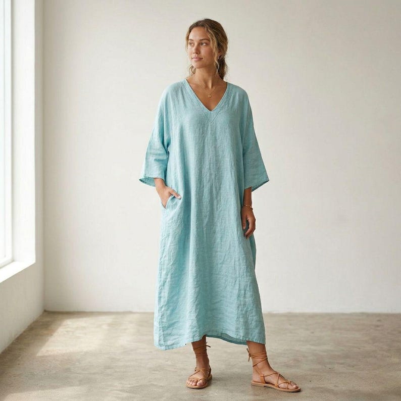 May include: A light blue, ankle-length dress with a V-neckline and three-quarter length sleeves. The garment features a loose, flowing silhouette with side pockets and a visible textured fabric finish, reaching down to the lower calf area.