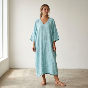 May include: A light blue, ankle-length linen dress with a V-neckline and loose, three-quarter length sleeves. The garment features a relaxed, straight-cut silhouette with a textured fabric finish, paired with brown strappy leather sandals.