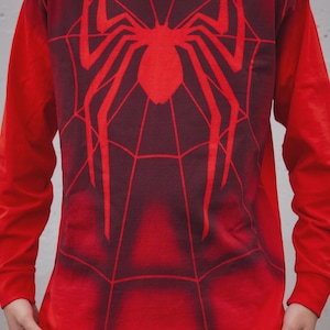 May include: A long-sleeve crew neck shirt in bright red featuring a dark red spider graphic on the chest. The design includes a web pattern that extends across the front of the garment, with a gradient effect transitioning from dark to light red.