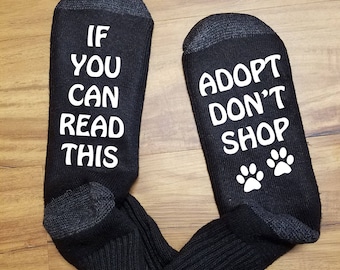 Op de afbeelding: Een paar zwarte sokken met witte tekst. Eén sok staat "Als je dit kunt lezen" en de andere sok staat "Adopteer, koop niet" met twee poot afdrukken.