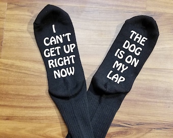 May include: A pair of black socks with white text. One sock reads "I can't get up right now" and the other reads "The dog is on my lap".