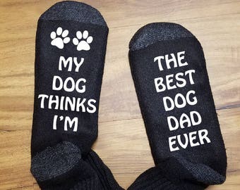 May include: A pair of black socks with white text. One sock reads "My dog thinks I'm" and the other sock reads "The best dog dad ever".