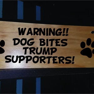 Puede incluir: Un letrero de madera con texto negro que dice "WARNING!! DOG BITES TRUMP SUPPORTERS!" con una huella de pata negra a cada lado.